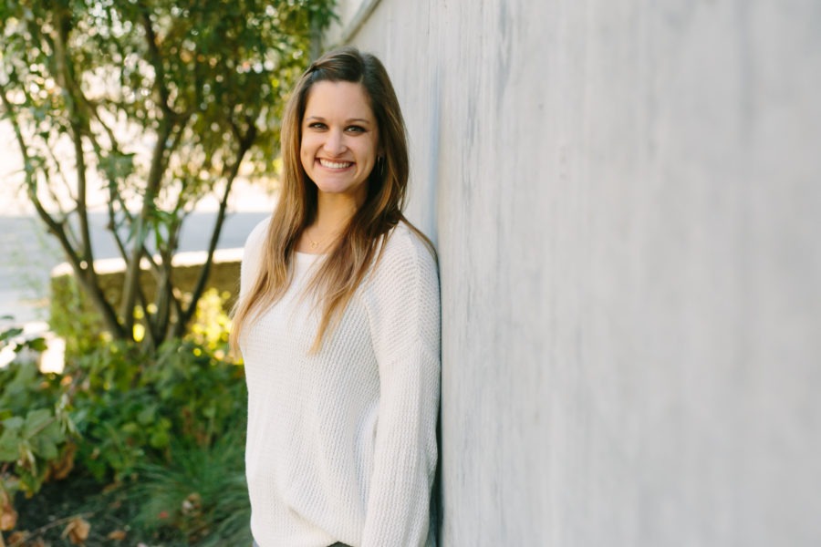 Author Katie Keridan stood in front of a wall with a tree and the sea in the background. She is wearing a white sweater and smiling