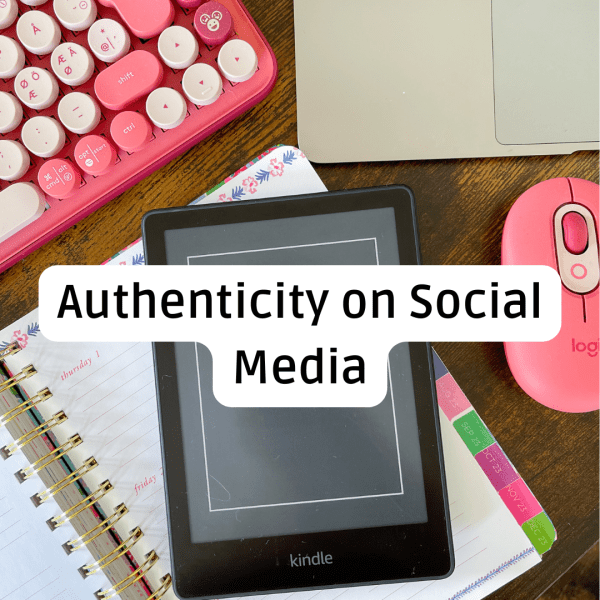 An overhead, flat-lay photograph of a desk setup featuring pink and white tech accessories on a dark wooden surface. A black Kindle e-reader is centered, resting on an open floral-patterned spiral planner. A large, white rounded text bubble is overlaid on the Kindle, reading "Authenticity on Social Media." Surrounding the center are several items: Top Left: A portion of a bright pink and white typewriter-style mechanical keyboard. Top Right: The corner of a silver laptop. Right Side: A vibrant pink Logitech pebble mouse. Bottom Right: Color-coded monthly tabs (Aug 23 through Dec 23) peeking out from the side of the planner.