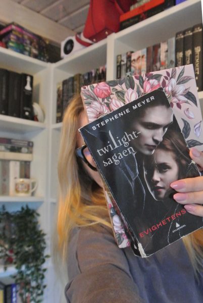 A person with long blonde hair and glasses holding two books in front of their face, partially obscuring it. The front book is a worn Norwegian edition of "Twilight" (titled "Twilight-sagaen: Evighetens kyss") featuring Edward and Bella on the cover. Behind it is a pink floral-patterned book. The background shows white bookshelves filled with various books.