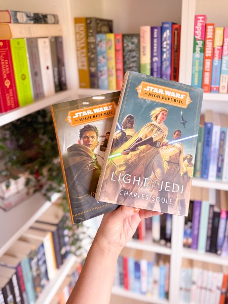 A hand holding two Star Wars books from The High Republic series, "Light of the Jedi" by Charles Soule and "Into the Dark" by Claudia Gray, in front of a white bookshelf filled with colorful novels.
