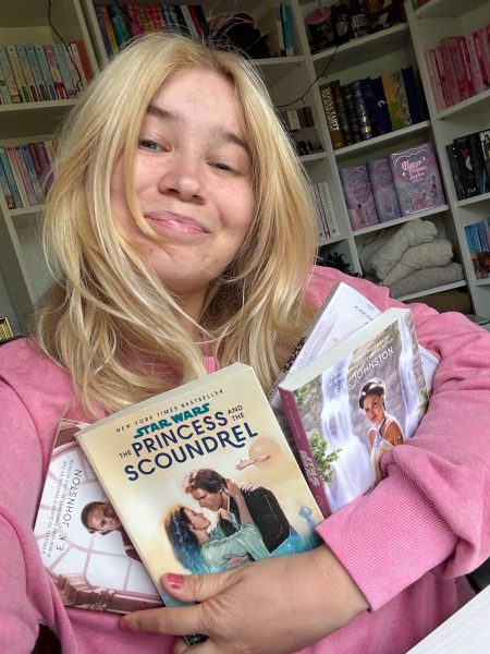 A person wearing a pink sweatshirt hugging a stack of Star Wars books, including "The Princess and the Scoundrel" and E.K. Johnston’s Queen’s Shadow series, in a cozy room with bookshelves.