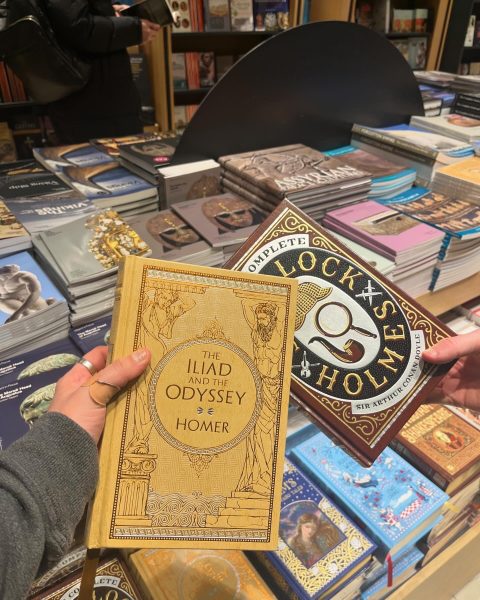 image of a hand holding a copy of The Iliad and the Odyssey by Homer. Behind is a table full of stacks of books